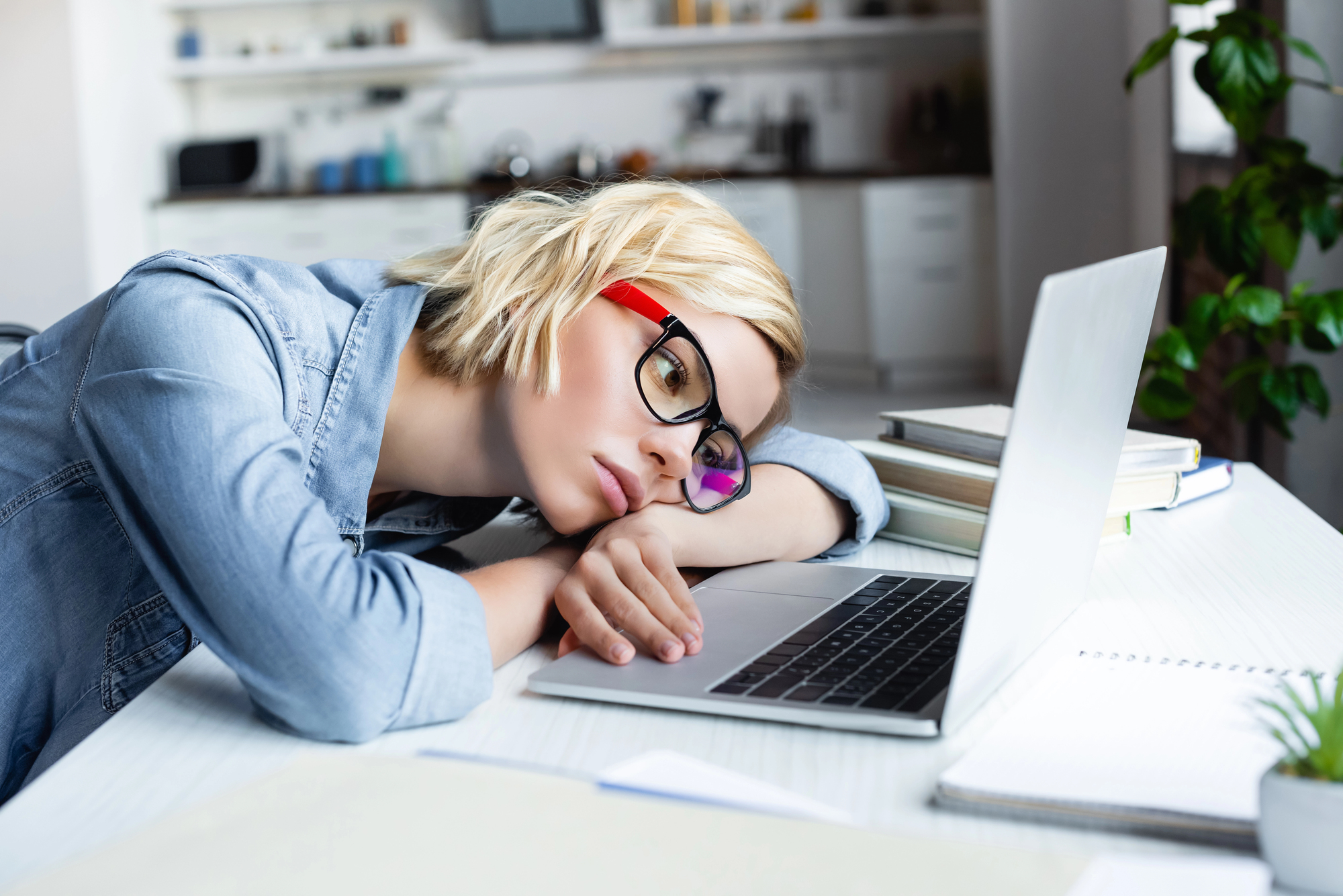 Tired blonde woman in eyeglasses with head on desk listlessly touching the touchpad of a laptop