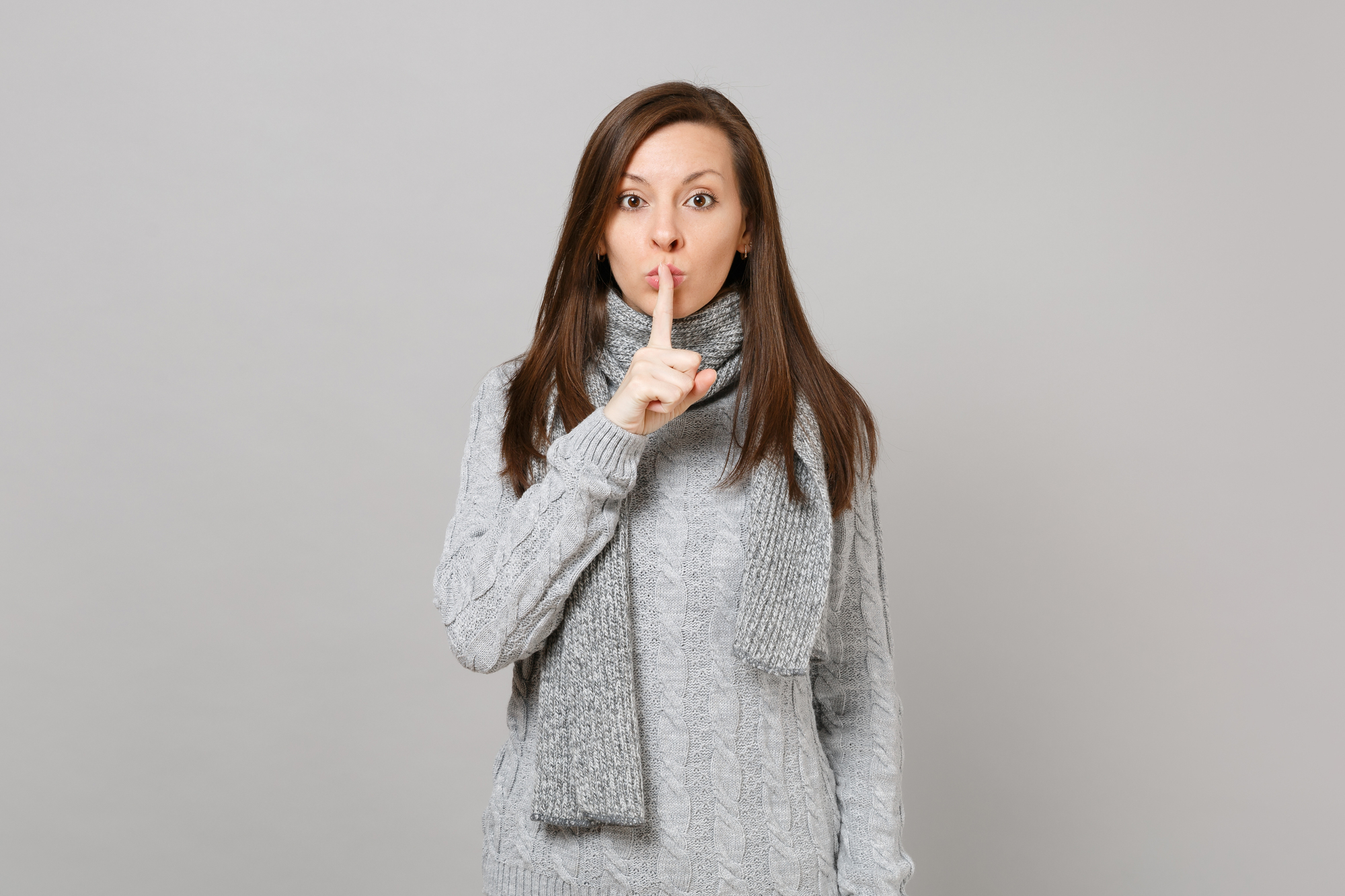 Young woman in gray sweater, scarf saying hush be quiet with finger on lips shhh gesture isolated on grey background.