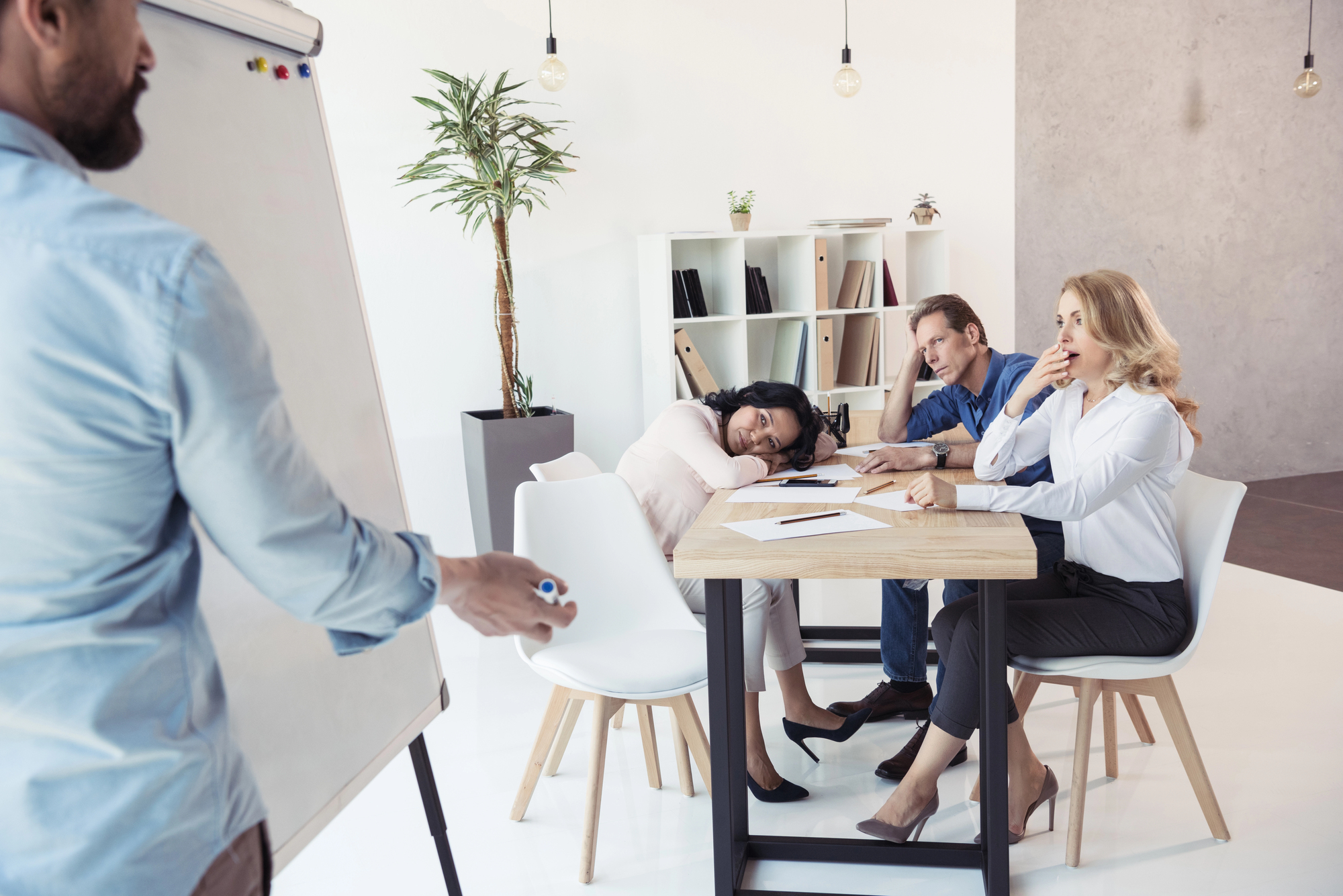 Bearded man presenting to bored colleagues sitting at a table across the room.